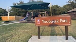 Uma área de recreação infantil com escorrega e estruturas de escalada, uma placa de madeira indicando 'Woodcreek Park' e uma área de estar coberta.