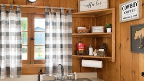 A kitchen with a sink, a coffee bar sign, a shelf with coffee-related items, and a window with curtains.