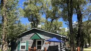 Uma cabana rústica com um alpendre de madeira, uma mesa e cadeiras, e um guarda-sol azul.