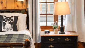 Een kamer met houten lambrisering, een bed, een nachtkastje met een lamp, een klok en een raam met gordijnen.
