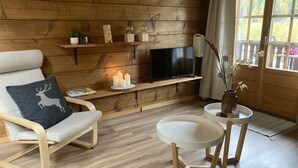 A cozy room with a wooden wall, a TV on a shelf, a chair with a cushion, a small table with a vase, and a window with a view of trees. - Luxury chalet with panoramic views in the Lötschental, with private carport  (Kippel)