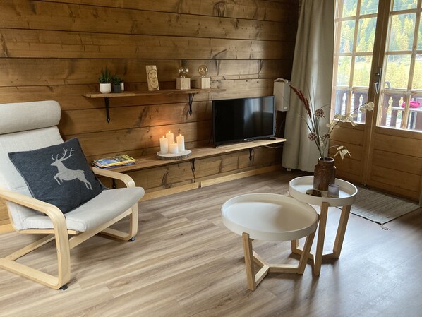 A cozy room with a wooden wall, a TV on a shelf, a chair with a cushion, a small table with a vase, and a window with a view of trees. - Luxury chalet with panoramic views in the Lötschental, with private carport (Kippel)