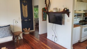 Cozinha moderna com armários brancos, forno embutido e micro-ondas. Há uma mesa de jantar de madeira com cadeiras, uma porta azul escura com um sonho apanhador e uma pequena área de estar com sofá e luminária.