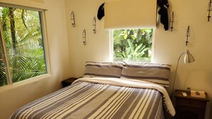 Cama bem arrumada com roupa de cama listrada, luminária de cabeceira, mesa de cabeceira e janela com vista para o verde.