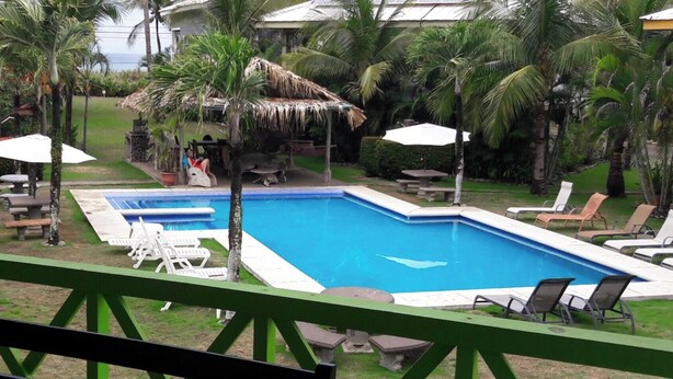 Piscina com espreguiçadeiras e guarda-sóis, cercada por palmeiras e uma estrutura de palha.