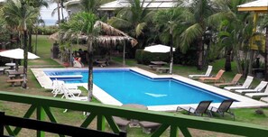 Una piscina amb gandules i ombrel·les, envoltada de palmeres i una construcció amb teulada de palla.