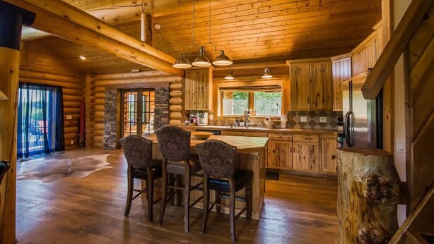 A log cabin interior with a central island, wooden dining chairs, a kitchen with a large window, and a staircase with a carved tree trunk. - Montana Wildflower Lodge~sleeps 10 (Red Lodge)