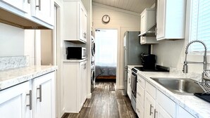 A narrow kitchen with white cabinets, a wooden floor, and a visible bed in the background.