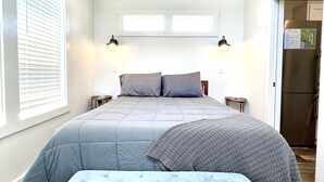 A bedroom with a bed, a bench, a ceiling fan, and a refrigerator.