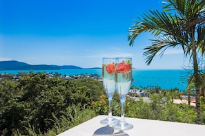 Two glasses of a clear beverage with fruit garnish, placed on a table with a view of a coastal landscape and a body of water.