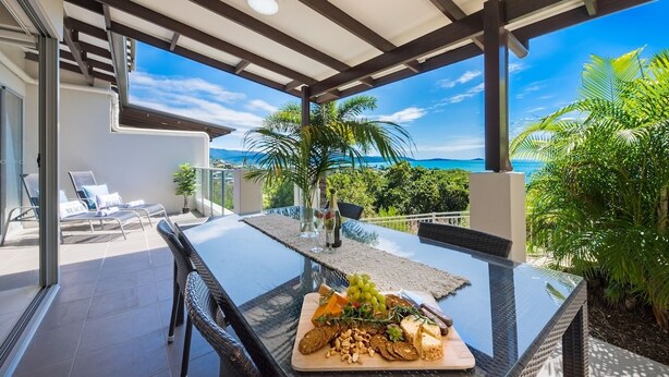 Mesa com tampo de vidro exibindo diversos queijos, frutas e pão, em varanda com vista para o oceano e palmeiras.