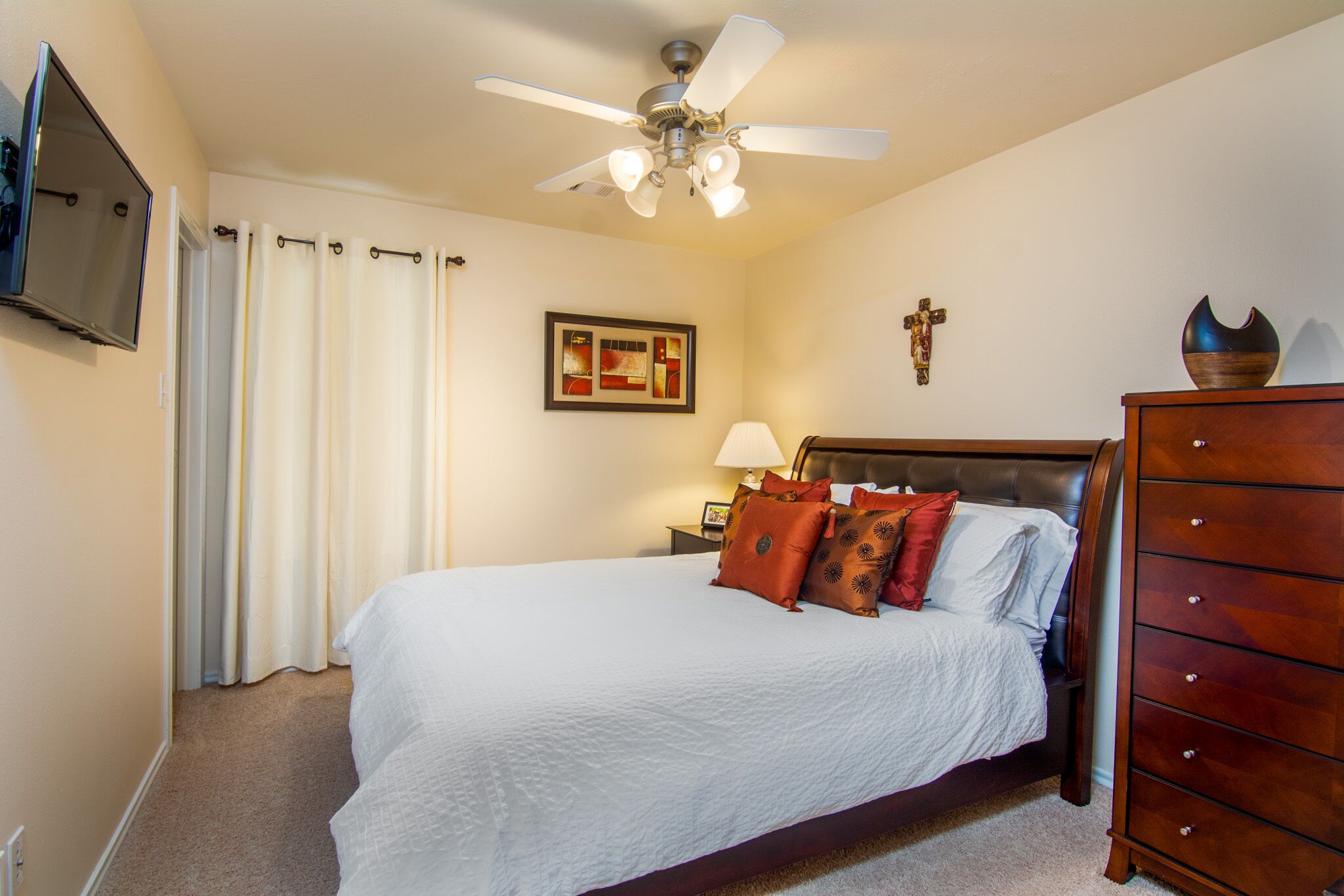 Une chambre à coucher avec un lit, un ventilateur de plafond, une table de chevet avec une lampe, une commode et un tableau encadré au mur.