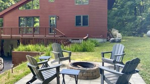 Una casa de dos pisos con un balcón, zona de estar exterior con sillas Adirondack y un brasero.