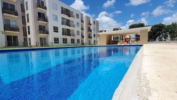 A swimming pool with a clear blue water surface, surrounded by a modern building with multiple balconies and a covered area with a slide.