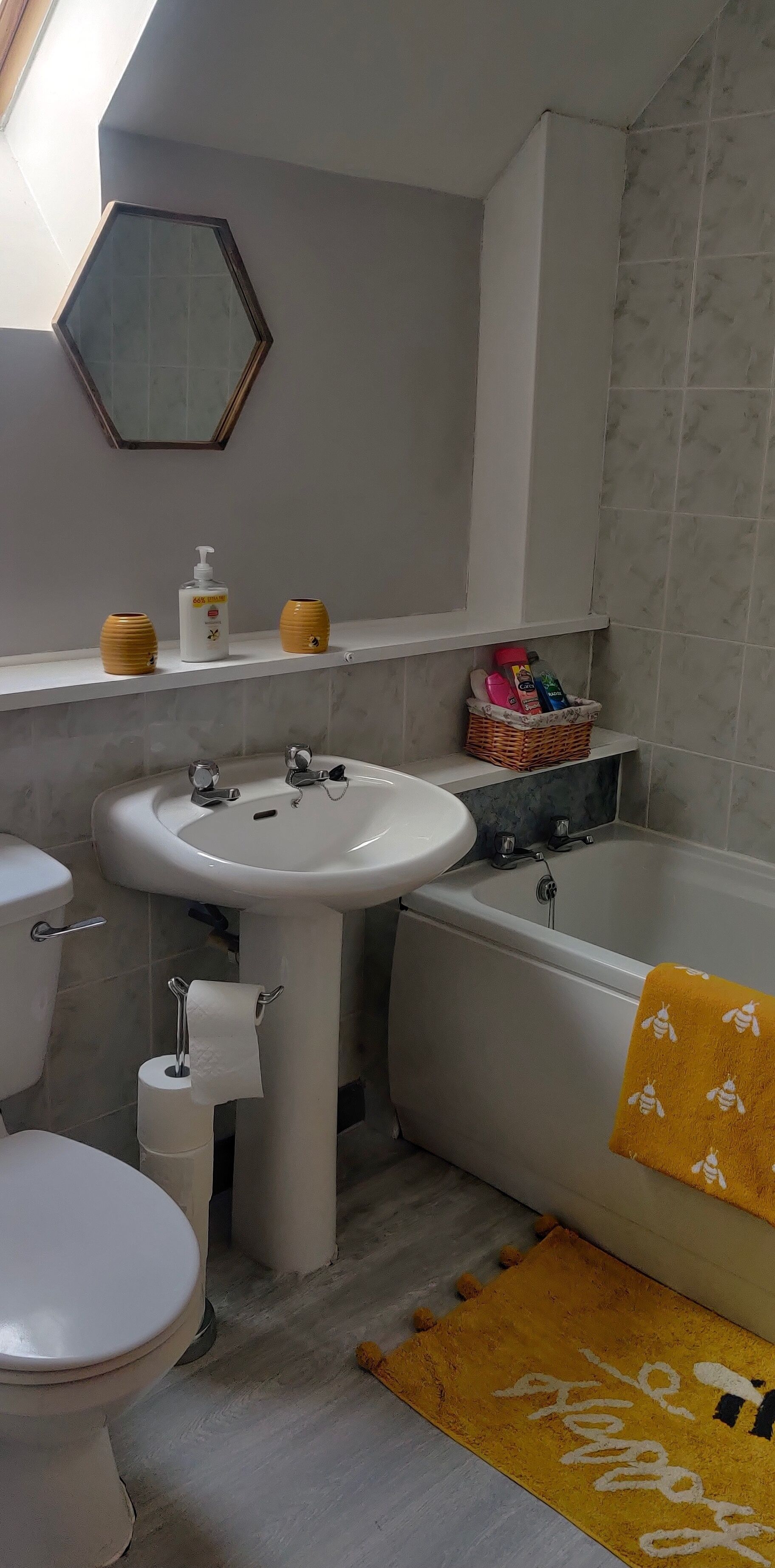 A bathroom with a sink, toilet, and bathtub. There are toiletries on the shelf and a yellow towel on the floor.
