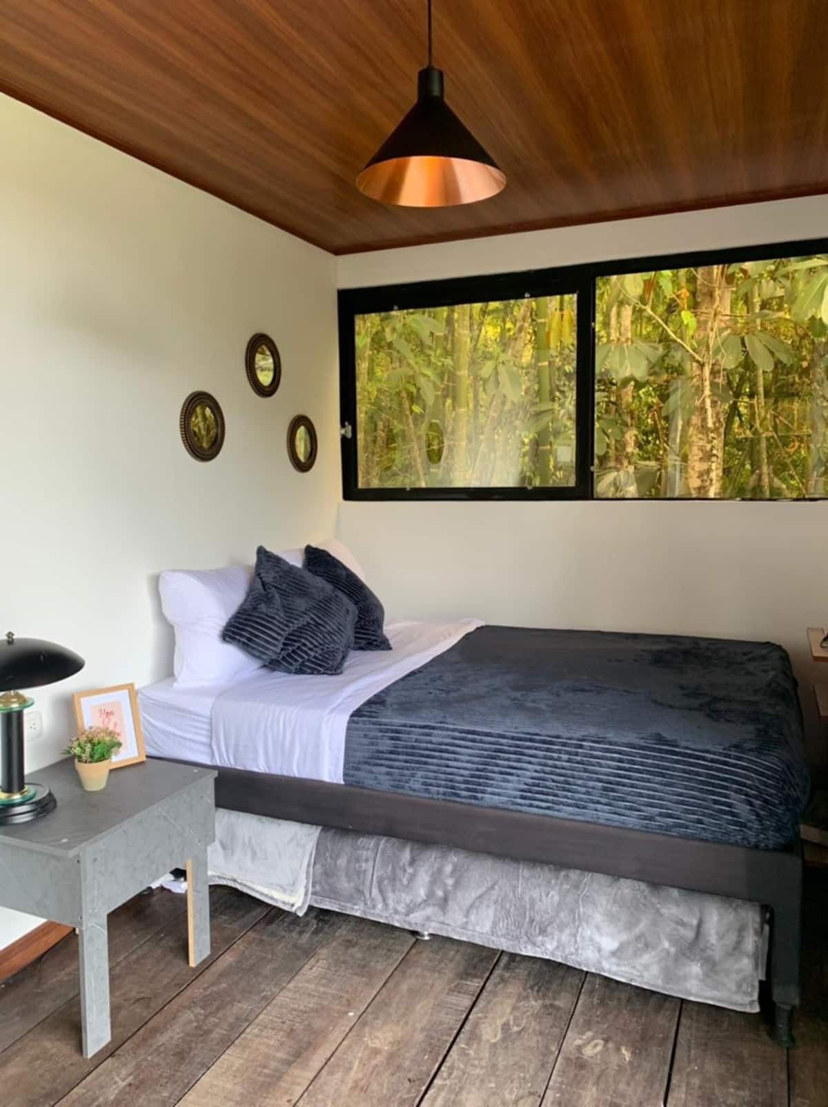 A bedroom with a bed, a desk with a lamp, a window with a view of trees, and a ceiling light.
