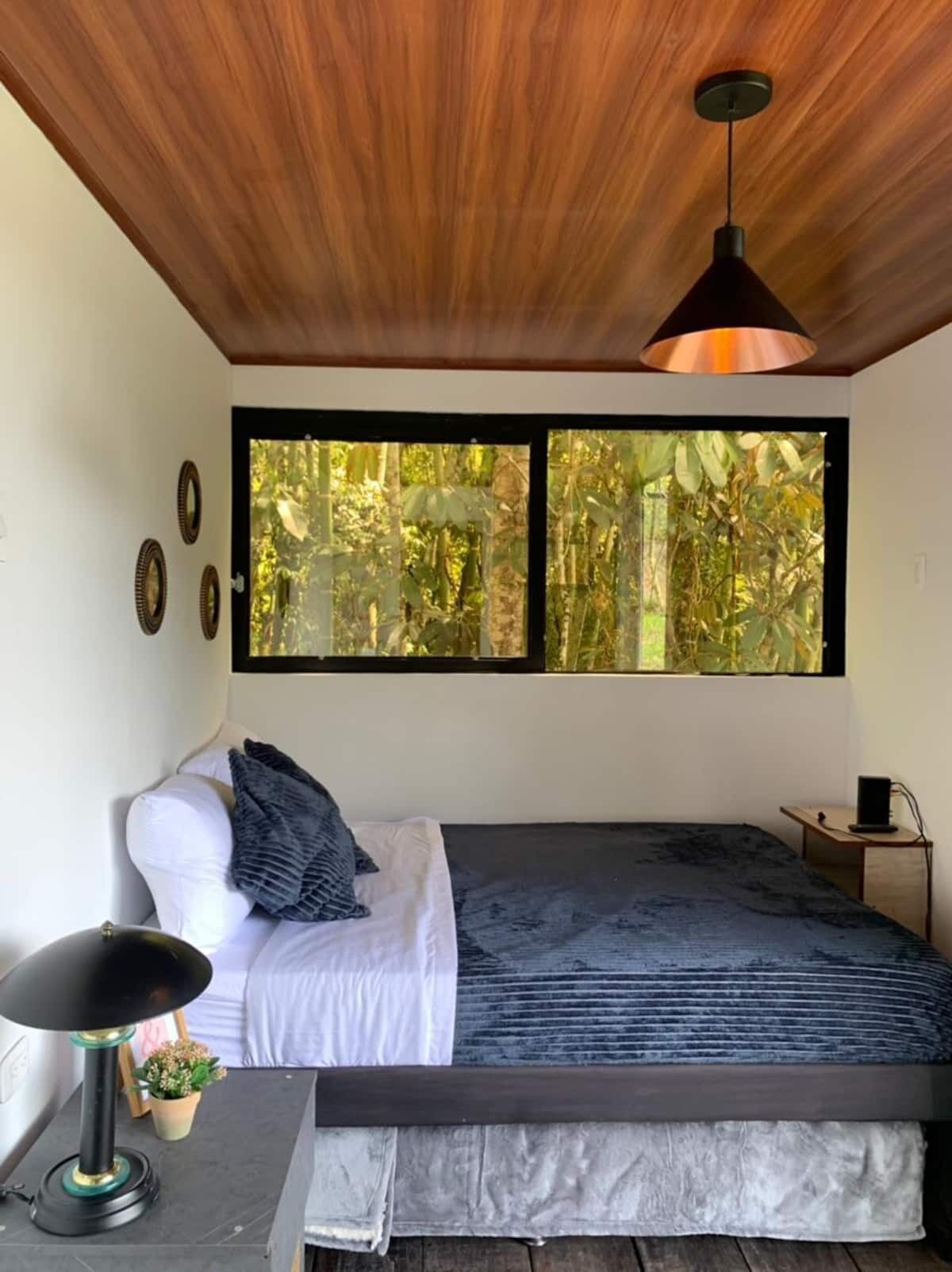 A bedroom with a bed, a desk, a lamp, and a window with a view of greenery.