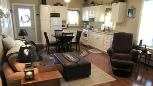 Ein Wohnzimmer mit Sofa, Hocker und einem kleinen Tisch mit Lampe. Es gibt einen Essbereich mit Tisch und Stühlen sowie eine Küche mit Schränken und Geräten.