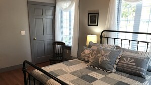 Ein Schlafzimmer mit einem Metallbettgestell, einer karierten Bettdecke und zwei Kissen mit Scheunen- und Sterndruck. Ein Stuhl, eine Tür und ein Fenster mit Jalousien sind ebenfalls vorhanden.