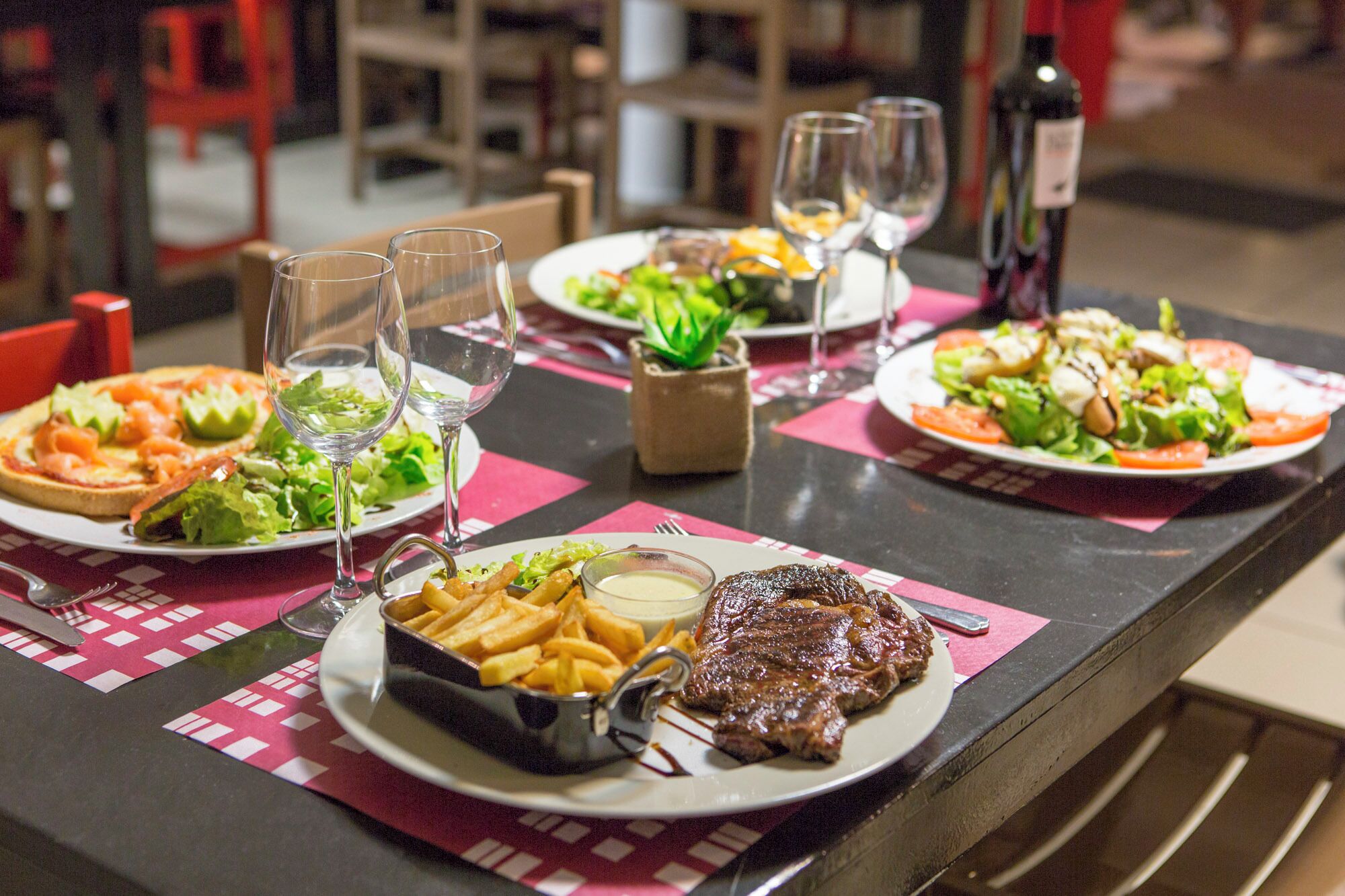 Une table à manger dressée avec divers plats, notamment un steak, des frites et des salades, ainsi que des verres à vin.
