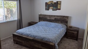 A bedroom with a wooden bed frame, a blue quilted bedspread, a nightstand with a clock, and a wall art piece.
