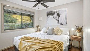 A bedroom with a large bed, a ceiling fan, a window with a view of greenery, and a mounted artwork of a horse.