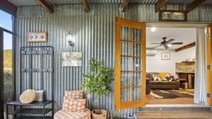 A rustic interior with a corrugated metal wall, a wooden door, a sofa with patterned cushions, a small table with a cushion, and a potted plant.