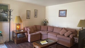 A living room with a brown leather sofa, a glass coffee table, and a wooden side table with a lamp. There are framed pictures on the wall and a vase with flowers on a side table.
