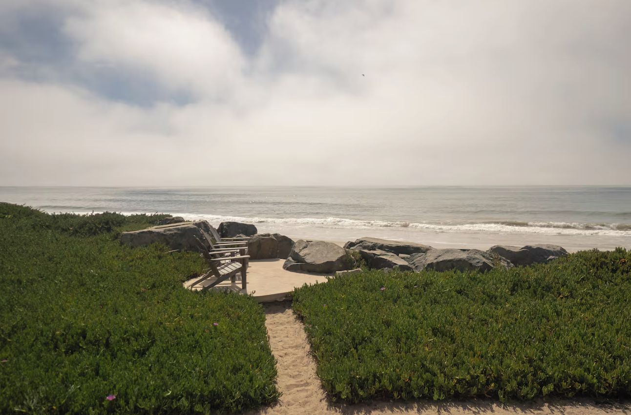 A beach path leading to a wooden bench overlooking the ocean, with large rocks and greenery in the foreground.