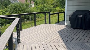 Una terraza de madera con barandilla, una parrilla y una casa al fondo.