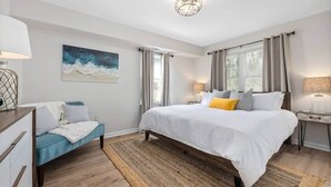 A bedroom with a large bed, a bedside table, a lamp, a window with curtains, a painting on the wall, and a wooden floor. - Stylish & Bright! Walk to Beach, Golf Cart, Patio (Isle of Palms)