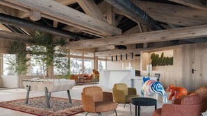 A spacious room with a high ceiling, wooden beams, and a large window overlooking a snowy landscape. There are several chairs and a small table, with a decorative sculpture on the right.
