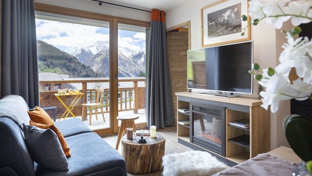 Ein modernes Wohnzimmer mit Sofa, Fernseher, Kamin und einem Balkon mit Blick auf die Berge.