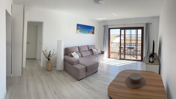 Un salon moderne avec un canapé gris, une table basse en bois et une vue sur un balcon aménagé avec du mobilier d’extérieur.
