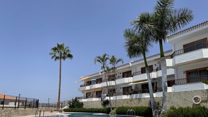 Une piscine entourée d’une terrasse carrelée, avec un immeuble à plusieurs étages doté de balcons et de palmiers en arrière-plan.