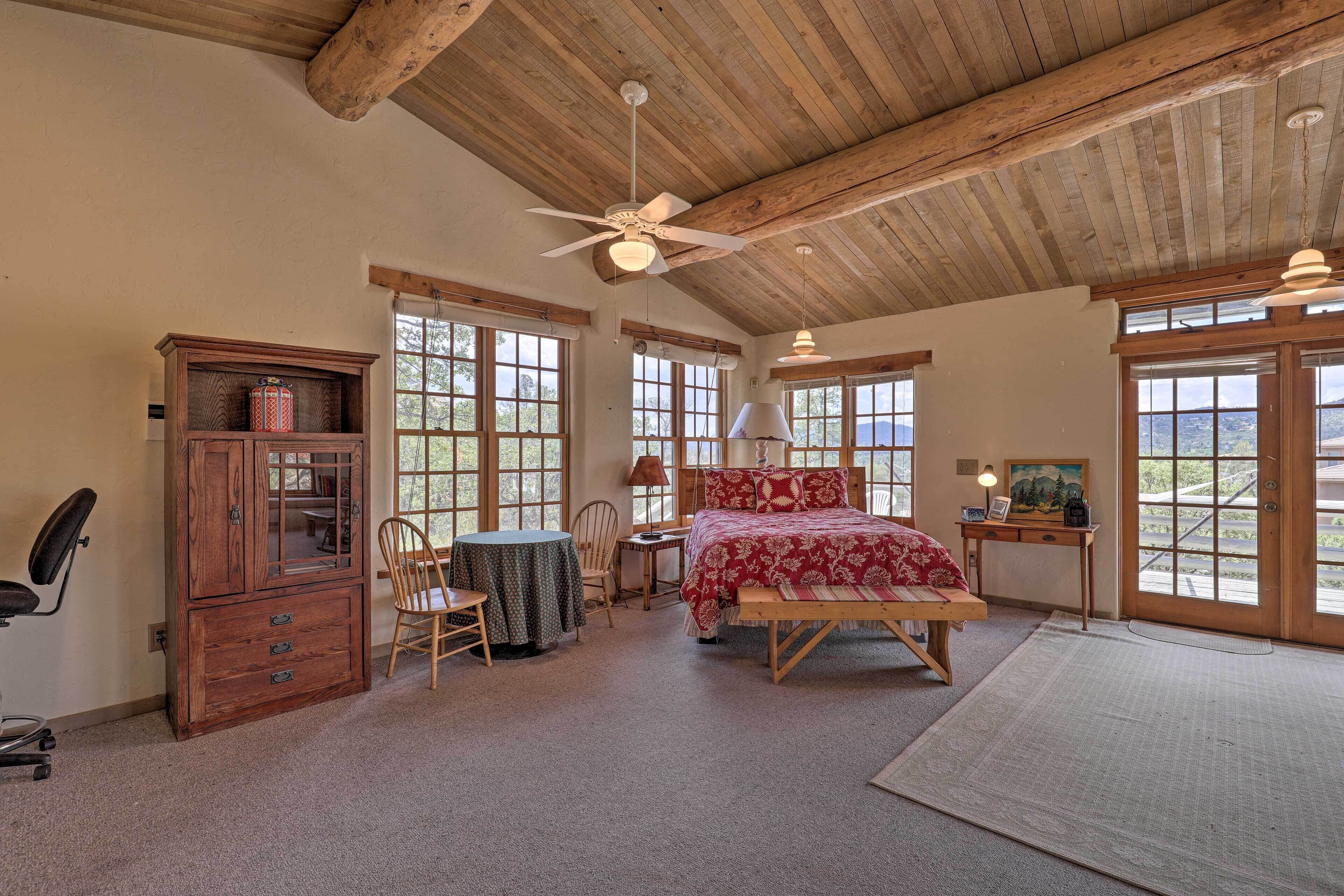 A bedroom with a wooden ceiling, a bed with red bedding, a desk with a lamp, and large windows.