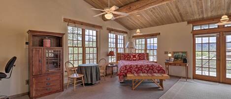 A bedroom with a wooden ceiling, a bed with red bedding, a desk with a lamp, and large windows.