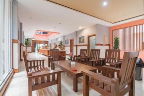 A spacious dining area with a long table and multiple chairs, a kitchen in the background, and a decorative wall with vertical elements. - Sans Hotel Budaya Cirebon (Kesambi)