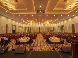 Un salón de conferencias con filas de mesas y sillas, piso alfombrado y un escenario al frente.