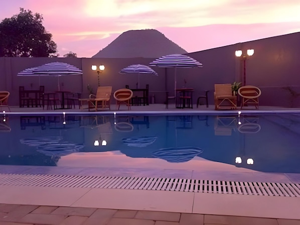 Una piscina con sillas de descanso y sombrillas, rodeada por un muro bajo y una montaña al fondo.