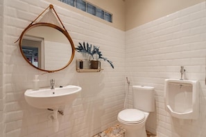 A bathroom with white tiled walls, a round mirror, a sink, a toilet, and a urinal. - Reddoorz Plus Bagusrangin Street (Bandung)