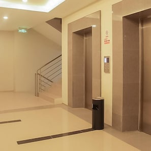 A modern elevator area with a handrail, emergency exit sign, and a trash bin. - Hotel Mutiara (Padang Sidempuan)