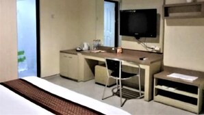 A hotel room with a bed, a desk with a chair, a television, and a small shelf. - Grand Mansion Hotel (Blitar)