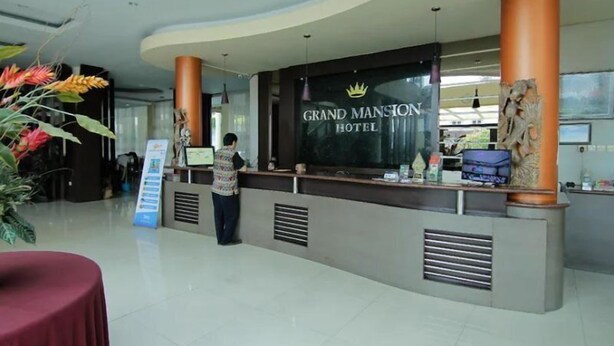 A reception desk at the Grand Mansion Hotel with a sign, a receptionist, and a potted plant. - Grand Mansion Hotel (Blitar)