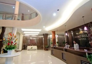 A modern hotel lobby with a reception desk, a seating area, and a staircase leading to an upper level. - Grand Mansion Hotel (Blitar)