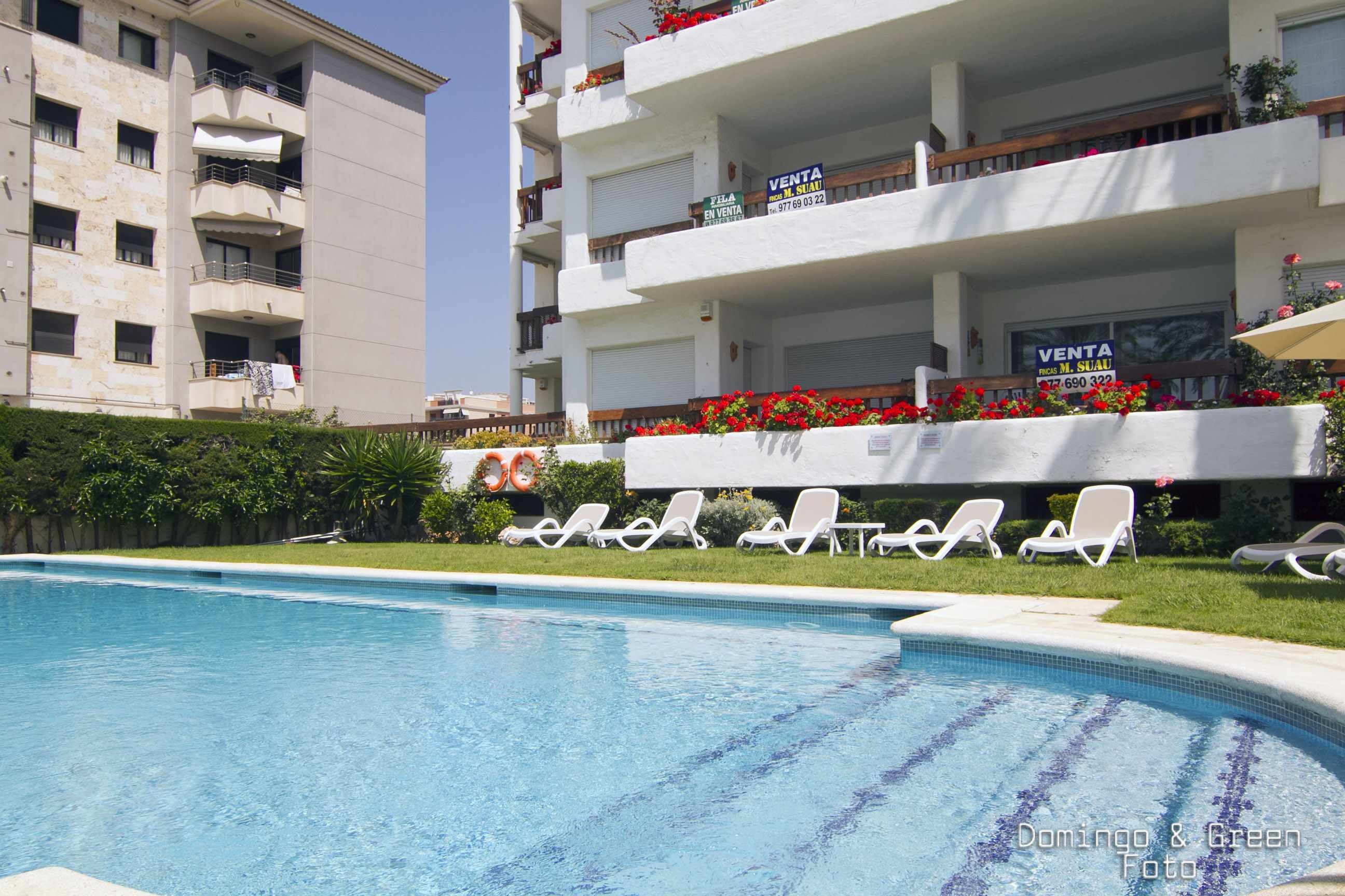 Une piscine avec des chaises longues et un immeuble doté de balcons et de panneaux indiquant les appartements disponibles à la location.