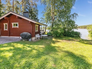 A red cabin with a covered grill, a patio table, and a chair, surrounded by a grassy yard and a lake. - Vacation home Elfvik by Interhome (Porvoo)