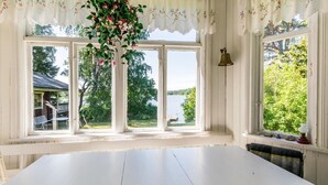 A room with a large white table, three windows with a view of a lake and greenery, and a hanging plant with pink flowers. - Vacation home Elfvik by Interhome (Porvoo)