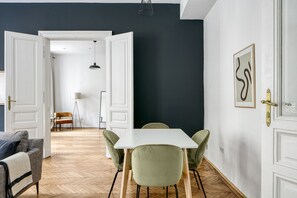 Uma sala de estar moderna com área de jantar, com uma mesa branca e cadeiras verdes, um sofá cinza e uma parede de destaque escura.