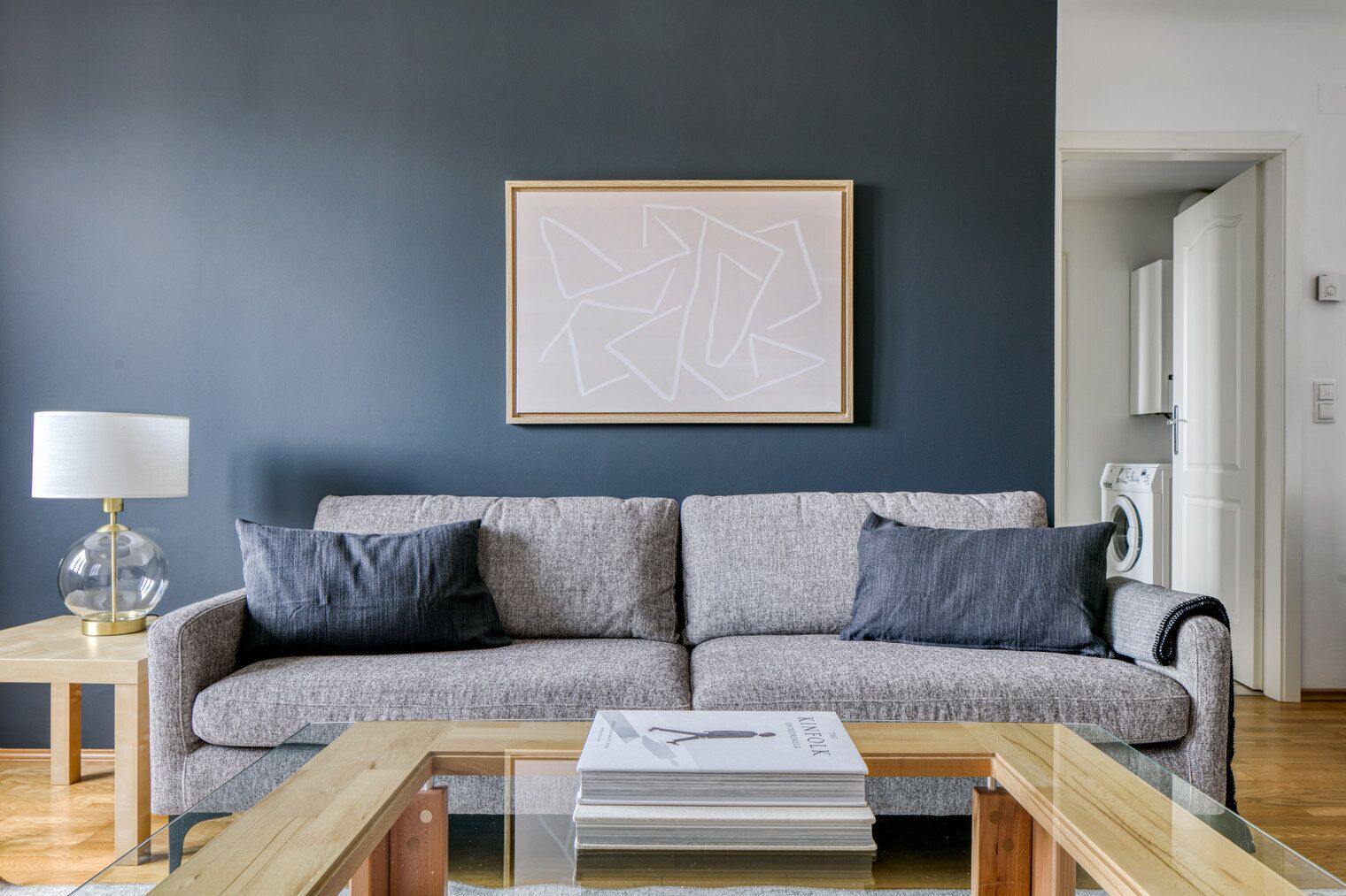 Una sala de estar moderna con un sofá gris, dos cojines oscuros, una mesa de centro de vidrio con un libro, una lámpara y una obra de arte abstracta enmarcada en la pared.